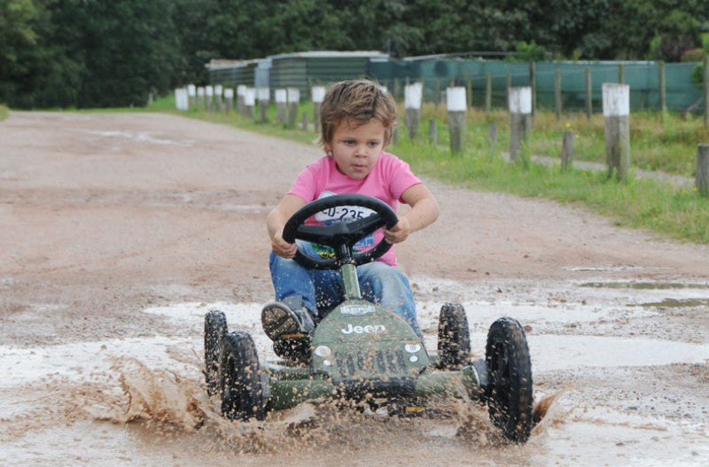 Liten pojke kör en pöl på en grusväg utanför i en Jeep Junior trampbil